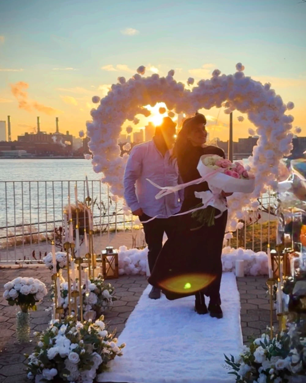Pareja abrazada bajo un arco de rosas blancas con el sol detrás